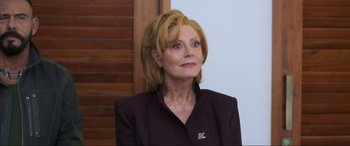 Movie still from “Blue Beetle” (2023), directed by Angel Manuel Soto – An older woman wearing a black suit and a brown jacket; Close Up shot, Low angle