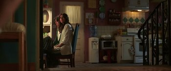 Movie still from “Blue Beetle” (2023), directed by Angel Manuel Soto – Two women sitting in a kitchen next to a microwave oven; Wide shot, Low angle