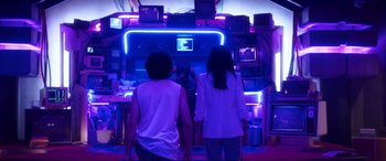 Movie still from “Blue Beetle” (2023), directed by Angel Manuel Soto – Two people standing in front of a television set; Wide shot, Low angle