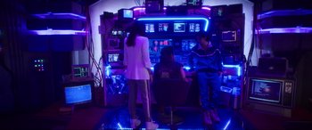 Movie still from “Blue Beetle” (2023), directed by Angel Manuel Soto – A group of people standing in front of an arcade machine; Wide shot, Over the shoulder angle