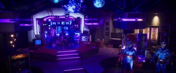Movie still from “Blue Beetle” (2023), directed by Angel Manuel Soto – People are sitting in a room with purple lighting; Extreme Wide shot, Low angle