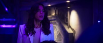 Movie still from “Blue Beetle” (2023), directed by Angel Manuel Soto – A woman in white jacket standing next to stairs; Medium shot, Over the shoulder angle
