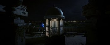Movie still from “Blue Beetle” (2023), directed by Angel Manuel Soto – A person sitting on a bench in front of a gazebo at night; Extreme Wide shot, High angle