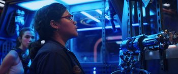 Movie still from “Blue Beetle” (2023), directed by Angel Manuel Soto – A woman with glasses is standing in front of a microphone; Medium shot, Low angle