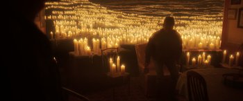 Movie still from “Blue Beetle” (2023), directed by Angel Manuel Soto – A person standing in front of many lit candles in a room; Extreme Wide shot, High angle