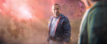 Movie still from “Blue Beetle” (2023), directed by Angel Manuel Soto – An older man standing in front of a colorful background; Medium shot, Low angle