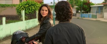 Movie still from “Blue Beetle” (2023), directed by Angel Manuel Soto – A woman in a black jacket is riding a bike; Medium shot, Over the shoulder angle