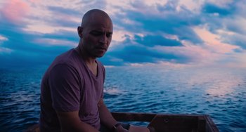 Movie still from “Blue Miracle” (2021), directed by Julio Quintana – A man sitting on top of a boat in front of a body of water; Medium shot, Low angle