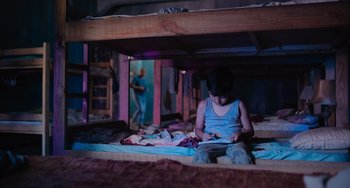 Movie still from “Blue Miracle” (2021), directed by Julio Quintana – A person sitting on a bed with a book; Wide shot, High angle