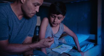 Movie still from “Blue Miracle” (2021), directed by Julio Quintana – A man and a boy sitting on a bed looking at a book; Medium shot, High angle