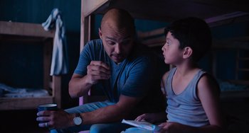 Movie still from “Blue Miracle” (2021), directed by Julio Quintana – A man sitting next to a boy holding a nintendo wii game controller; Medium shot, Over the shoulder angle