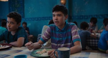 Movie still from “Blue Miracle” (2021), directed by Julio Quintana – A young man sitting at a table eating a plate of food; Medium shot, Over the shoulder angle