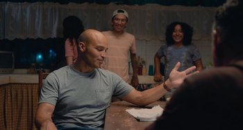 Movie still from “Blue Miracle” (2021), directed by Julio Quintana – A man sitting at a table in front of a group of people; Medium shot, Over the shoulder angle