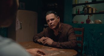 Movie still from “Blue Miracle” (2021), directed by Julio Quintana – A man sitting at a wooden table in a room; Medium shot, Over the shoulder angle