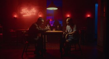 Movie still from “Blue Miracle” (2021), directed by Julio Quintana – Two men sitting at a table in front of a neon sign; Wide shot, Over the shoulder angle