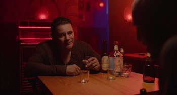 Movie still from “Blue Miracle” (2021), directed by Julio Quintana – A man sitting at a table in front of a bottle of alcohol; Medium shot, Over the shoulder angle