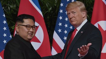Movie still from “Borat Subsequent Moviefilm” (2020), directed by Jason Woliner – A man in a suit standing next to another man in a suit; Medium shot, Over the shoulder angle