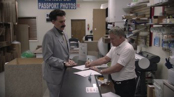 Movie still from “Borat Subsequent Moviefilm” (2020), directed by Jason Woliner – A man signing a document at a counter while another man looks on; Medium shot, Low angle