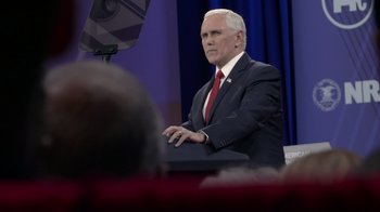 Movie still from “Borat Subsequent Moviefilm” (2020), directed by Jason Woliner – A man wearing a suit and tie standing in front of a microphone; Medium shot, Over the shoulder angle