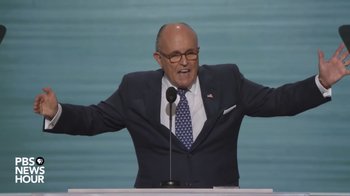 Movie still from “Borat Subsequent Moviefilm” (2020), directed by Jason Woliner – A man in a suit and tie standing at a podium; Medium shot, Low angle