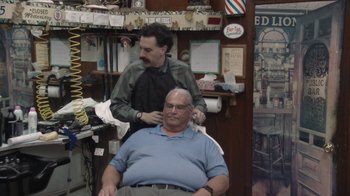 Movie still from “Borat Subsequent Moviefilm” (2020), directed by Jason Woliner – A man sitting in front of another man in a barber shop; Medium shot, Over the shoulder angle