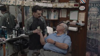 Movie still from “Borat Subsequent Moviefilm” (2020), directed by Jason Woliner – An older man sitting in a barber chair while a barber gives him money; Medium shot, Over the shoulder angle
