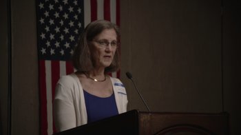 Movie still from “Borat Subsequent Moviefilm” (2020), directed by Jason Woliner – A woman standing in front of a microphone in front of an american flag; Medium shot, Over the shoulder angle