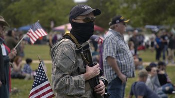 Movie still from “Borat Subsequent Moviefilm” (2020), directed by Jason Woliner – A man in camouflage holding a rifle in a crowd; Medium shot, Over the shoulder angle