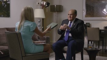 Movie still from “Borat Subsequent Moviefilm” (2020), directed by Jason Woliner – A man in a suit sitting in front of a woman in a blue dress; Medium shot, Over the shoulder angle