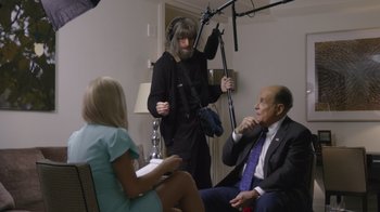 Movie still from “Borat Subsequent Moviefilm” (2020), directed by Jason Woliner – Two men and a woman are sitting in front of a microphone; Medium shot, Over the shoulder angle