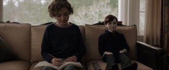 Movie still from “Brahms: The Boy II” (2020), directed by William Brent Bell – Two young children sitting on a couch and one of them is reading a book; Medium shot, Over the shoulder angle