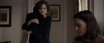 Movie still from “Brahms: The Boy II” (2020), directed by William Brent Bell – A young woman hugging a stuffed animal while standing in a living room; Medium shot, Over the shoulder angle