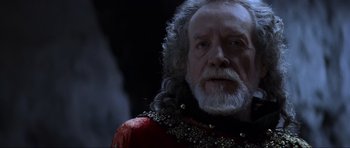 Movie still from “Braveheart” (1995), directed by Mel Gibson – An old man with a beard and a long white beard wearing a red and black outfit; Close Up shot, Over the shoulder angle