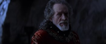 Movie still from “Braveheart” (1995), directed by Mel Gibson – An old man with a beard and long grey hair wearing a red and black outfit; Close Up shot, Over the shoulder angle