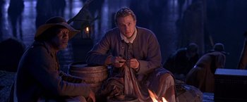 Movie still from “The Patriot” (2000), directed by Roland Emmerich – A man sitting on the ground in front of a fire; Medium shot, Over the shoulder angle