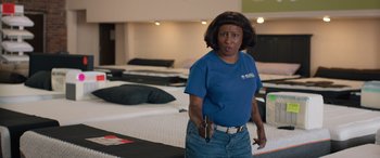 Movie still from “Breaking News in Yuba County” (2021), directed by Tate Taylor – A woman holding a cell phone standing in a room full of mattresses; Medium shot, High angle