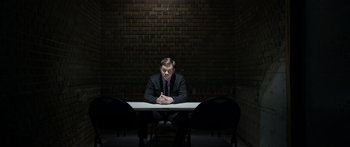 Movie still from “Broken” (2021), directed by Patrick Phillips – A man sitting at a table in front of a brick wall; Wide shot, High angle