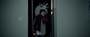 Movie still from “Broken” (2021), directed by Patrick Phillips – A young woman standing in front of a door wearing winter clothing; Medium shot, Over the shoulder angle
