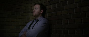 Movie still from “Broken” (2021), directed by Patrick Phillips – A man standing in front of a brick wall with his arms crossed; Medium shot, Low angle