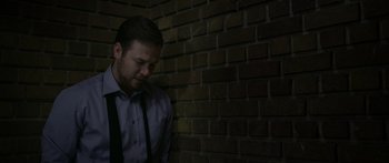 Movie still from “Broken” (2021), directed by Patrick Phillips – A man wearing a dress shirt and a tie leaning up against a brick wall; Medium shot, Low angle