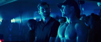Movie still from “Bros” (2022), directed by Nicholas Stoller – A man standing next to another man in a dark room; Medium shot, Over the shoulder angle