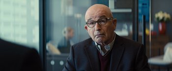 Movie still from “Bros” (2022), directed by Nicholas Stoller – An older bald man wearing glasses and a jacket; Close Up shot, Over the shoulder angle