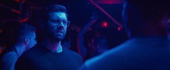 Movie still from “Bros” (2022), directed by Nicholas Stoller – A man with glasses is standing in a dark room; Close Up shot, Over the shoulder angle