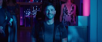Movie still from “Bros” (2022), directed by Nicholas Stoller – A young man smiles brightly at the camera in a dark room; Close Up shot, Over the shoulder angle