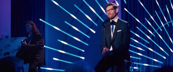 Movie still from “Bros” (2022), directed by Nicholas Stoller – A man standing in front of a microphone on a stage; Medium shot, Over the shoulder angle