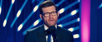 Movie still from “Bros” (2022), directed by Nicholas Stoller – A man in a suit and tie is speaking into a microphone; Close Up shot, Over the shoulder angle