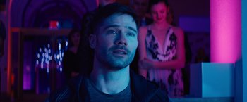 Movie still from “Bros” (2022), directed by Nicholas Stoller – A man in a dark room with people in the background; Close Up shot, Over the shoulder angle