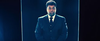 Movie still from “Bros” (2022), directed by Nicholas Stoller – A man wearing a suit and tie standing in front of a black background; Medium shot, Low angle