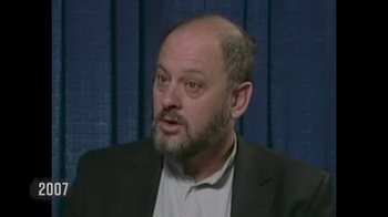 Movie still from “Burning” (2021), directed by Eva Orner – A man with a beard and a suit and tie talking; Close Up shot, High angle