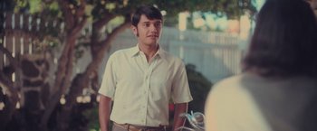 Movie still from “Burning Patience” (2022), directed by Rodrigo Sepúlveda – A man in a white shirt and brown pants standing in front of a tree; Medium shot, Over the shoulder angle